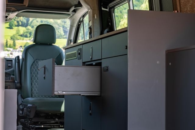 Innenausbau von Cocoon Vans eines VW Crafters 4x4 mit Holzakzenten aus amerikanischem Nussbraum, zwei grünen Ledersitzen, viel Stauraum über Kopf und einem kompakten Küchenbereich. Die zweite Schublade der Küche ist geöffnet.