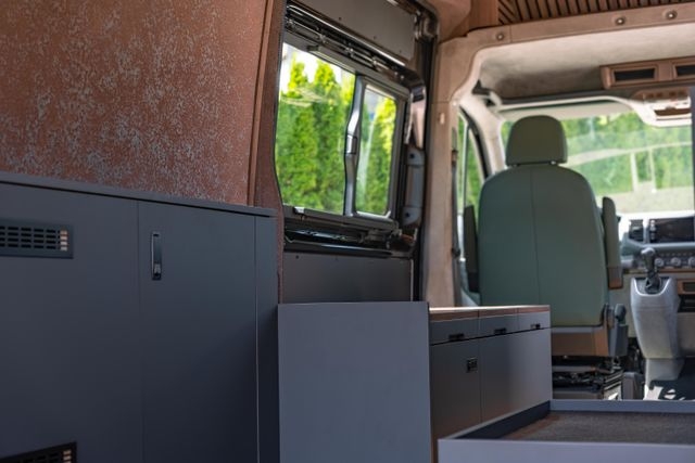 Innenansicht eines VW Crafters 4x4 von Cocoon Vans mit Stauschränken, Möbeln mit viel Stauraum und einer Seitentüre mit einem grossen Fenster, einer Wandbeplankung aus Leichtbauplatte aus Rostimitat die leicht magnnetisch ist. Der Fahrersitz ist vorne zu sehen.