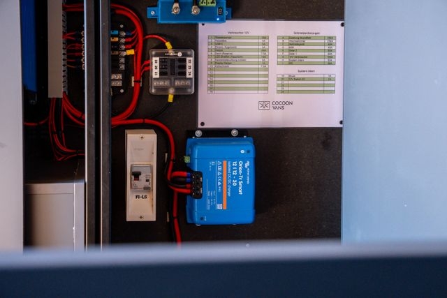 Elektronik eingebaut von Cocoon Vans. Eine Schalttafel mit Kabeln, blauen Victron Energy-Geräten, Schaltern und einer Checkliste, die auf einer schwarzen Tafel angebracht ist.